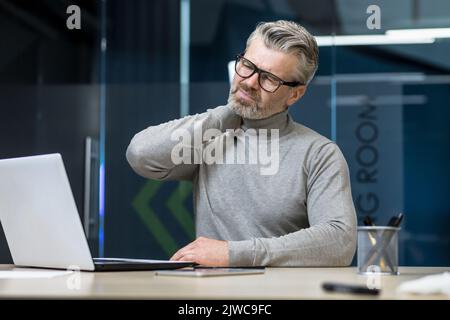 Senior mature businessman working inside office building, gray haired man sick and tired massaging neck with hand, boss having severe pain in neck and back. Stock Photo