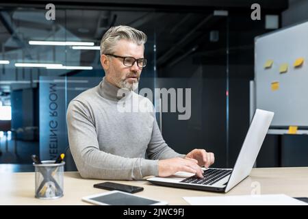 Senior and experienced programmer works in modern office at laptop, male web designer developer in glasses writes code programs new application, mature businessman works at desk. Stock Photo