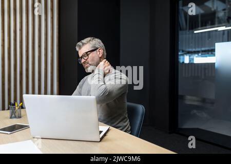 Senior mature businessman working inside office building, gray haired man sick and tired massaging neck with hand, boss having severe pain in neck and back. Stock Photo