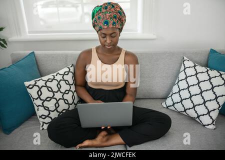Smiling young African American woman sitting with her legs crossed on her sofa at home using laptop Stock Photo