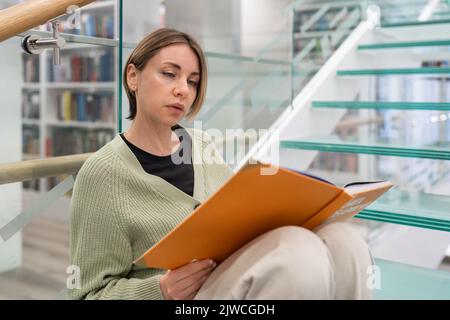 Focused female student enjoys reading interesting book with orange cover sitting on glass stairs Stock Photo