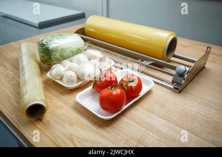 Close up of vegetables on white plastic trays lie on table near roll of stretch film. Use of food polyethylene transparent film for food storage in re Stock Photo