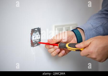 Man doing electrics. Close up of man installing electrical outlet in wall in room of house. Male electrician uses screwdriver to tighten bolts while a Stock Photo