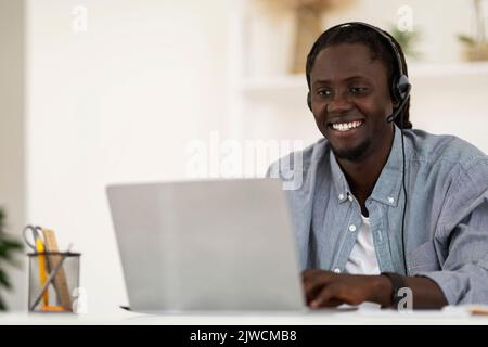Telecommuting Concept. Smiling African American Man Wearing Headset Working With Laptop Computer Stock Photo