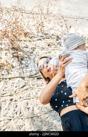 Mother in sunglasses holds cute toddler in arms against the wall. Young brunette woman and little boy enjoy summer vacation low angle close view Stock Photo