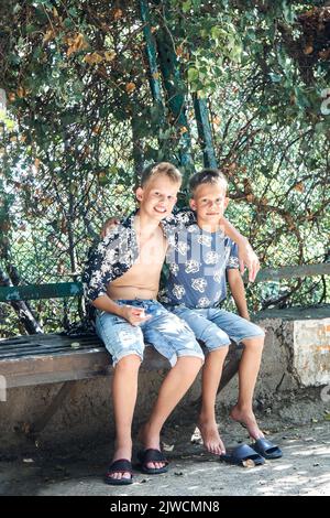 Friendly siblings sit on stone bench resting in tree shadow on summer holidays. Two blond brothers in shorts enjoy spending time together on sunny day Stock Photo