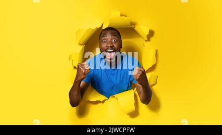 Overjoyed black man shouting happily, clenching both fists, cheering great success, posing in ripped yellow paper hole Stock Photo