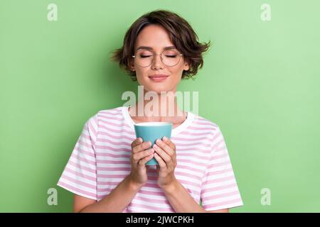 Photo of adorable cute lady dressed stylish striped outfit eyes closed hold hot tea enjoy free time isolated on green color background Stock Photo