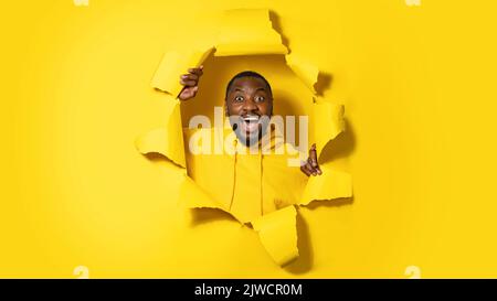 Excited african american man peeking out of hole in torn yellow wall background, banner, copy space Stock Photo