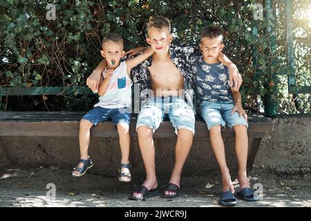 Friendly siblings sit on stone bench resting in tree shadow on summer holidays. Three blond brothers in shorts enjoy spending time together Stock Photo