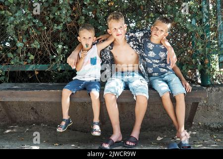 Friendly siblings sit on stone bench resting in tree shadow on summer holidays. Three blond brothers in shorts enjoy spending time together Stock Photo