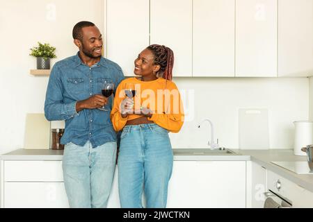 Loving black couple drinking red wine at kitchen, happy man and woman standing with glasses, copy space Stock Photo