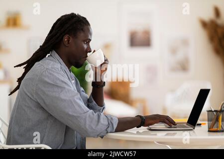 Remote Work. Black Freelancer Guy Working With Laptop And Drinking Coffee Stock Photo