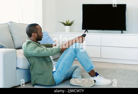 African american man watching television, pointing remote control at empty screen, mockup Stock Photo