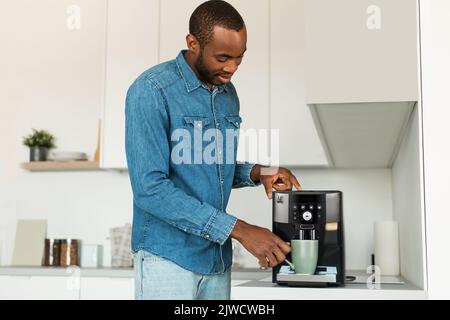 Handsome young african american man using modern coffee machine in kitchen, making fresh aromatic coffee Stock Photo