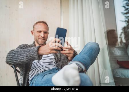 Smiling middle-aged man dressed open cardigan, jeans, and warm socks sitting on house balcony and smiling when he using modern smartphone. Everyday li Stock Photo