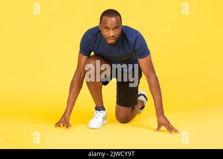 Black Male Runner Standing In Crouch Start Position, Yellow Background Stock Photo