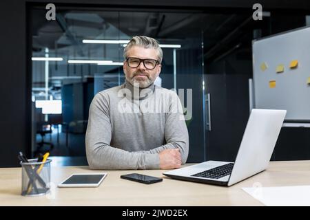 Portrait of senior programmer, mature team leader serious and focused looking at camera, businessman boss working inside office building, using laptop. Stock Photo