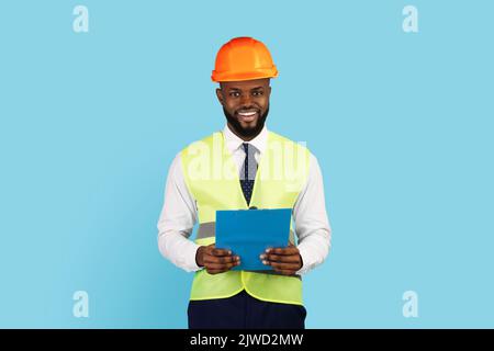 Professions Concept. Handsome Black Engineer In Hard Hat And Vest Holding Clipboard Stock Photo