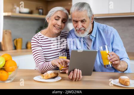 Cheerful Elderly Couple Having Breakfast Together And Using Digital Tablet In Kitchen Stock Photo
