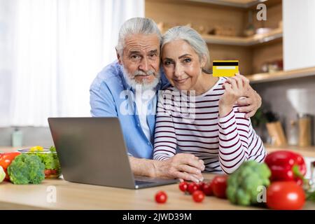 E-Commerce Concept. Senior Couple Holding Credit Card And Using Laptop In Kitchen Stock Photo