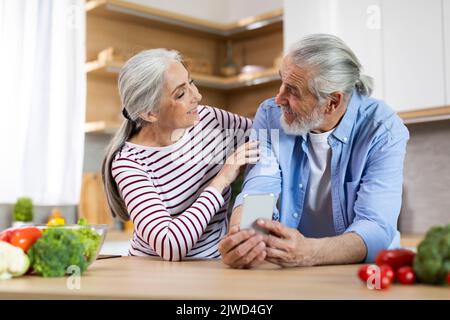 Modern Technologies. Happy Senior Spouses Using Smartphone While Relaxing In Kitchen Together Stock Photo