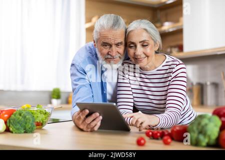 Portrait Of Happy Elderly Spouses Using Digital Tablet In Kitchen Interior, Stock Photo