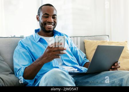 Business and telecommuting concept. Young black man using smartphone, sitting on sofa with laptop computer Stock Photo