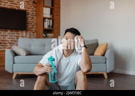 After training. Tired asian man with white towel on neck holding shaker with water and swiping sweat from forehead Stock Photo