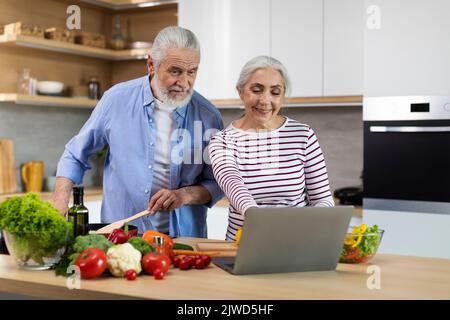 Cheerful Elderly Spouses Looking Recipe On Laptop While Cooking Food In Kitchen Stock Photo