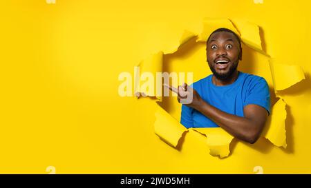 Excited black man gazing at camera with happy expression, pointing at free space, peeking through yellow background Stock Photo