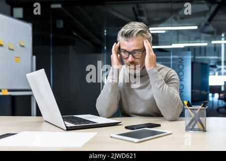 Sick mature businessman working in modern office, senior experienced man having severe headache, manager massaging temples while sitting on chair, manager using laptop for work Stock Photo