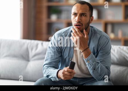 Shocked Black Man With Remote Controller In Hand Sitting On Couch Stock Photo