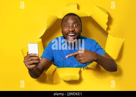 Shocked african american man holding cellphone and pointing on it, standing through yellow paper torn hole Stock Photo
