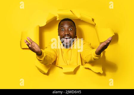 Excited african american man spreading hands sideways, looking at camera with open mouth through yellow paper hole Stock Photo