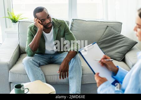 Depressed black man suffering from nevrous breakdown, having session at psychologist's office, sitting on sofa Stock Photo