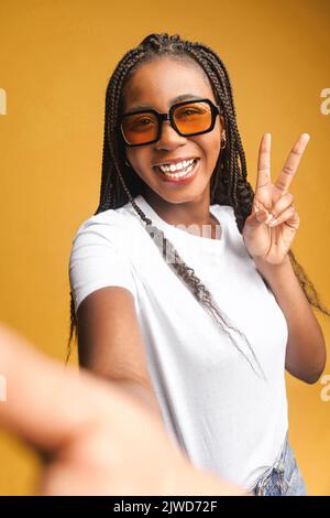 Happy carefree African-American young woman taking selfie on the smartphone, gesturing, making content for social medias, isolated on yellow Stock Photo