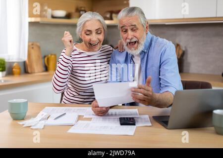 Portrait of excited senior spouses with papers celebrating good news in kitchen Stock Photo