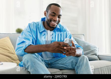 Excited black man chatting on smartphone, watching video, playing game while resting on sofa in living room Stock Photo