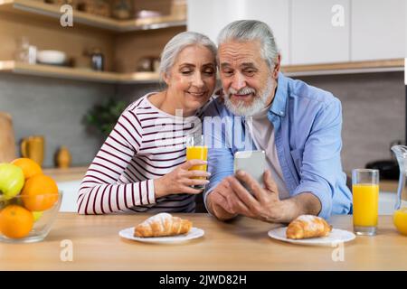 Portrait Of Happy Senior Spouses Using Smartphone During Breakfast In Kitchen Stock Photo