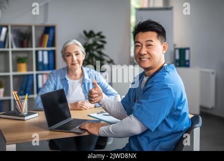 Cheerful adult asian male doctor showing thumb up sign with hand and consultation elderly female patient Stock Photo