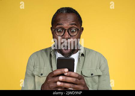 Shocked happy adult african american male in casual with open mouth in glasses looks at smartphone Stock Photo