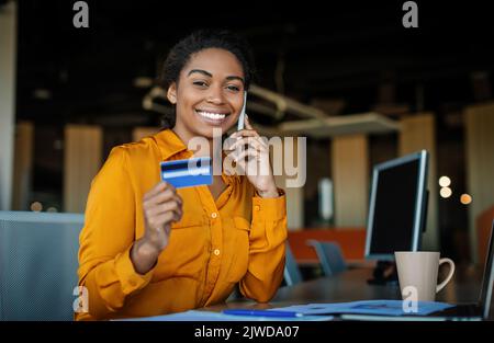 Easy payments concept. Happy black female worker holding credit card after online purchase and talking on smartphone Stock Photo