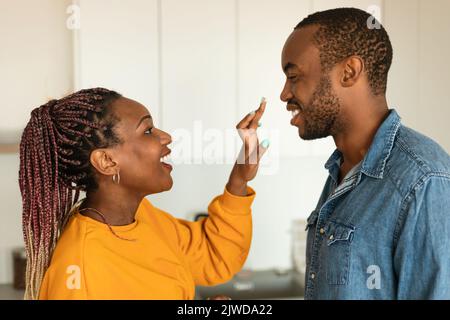 Happy african american spouses baking pie and having fun in kitchen, woman smearing husband's nose with flour Stock Photo