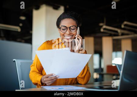 Happy black businesswoman talking on cellphone and checking documents in office, working with papers at workplace Stock Photo
