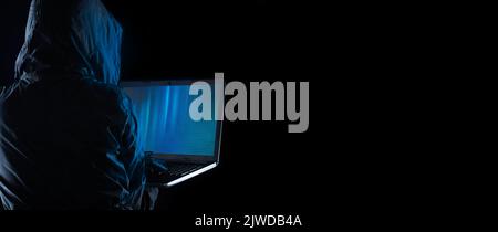Hooded cybercriminal uses laptop to hack internet in cyberspace, black background, internet personal data security concept. Hacker on a black backgrou Stock Photo