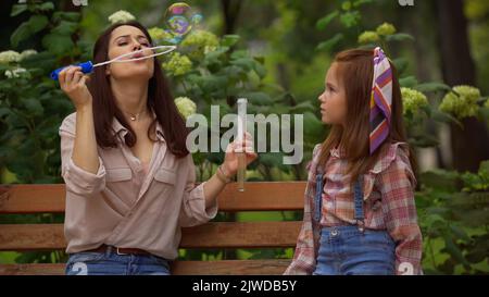 Woman blowing soap bubbles near redhead daughter on bench in park,stock image Stock Photo