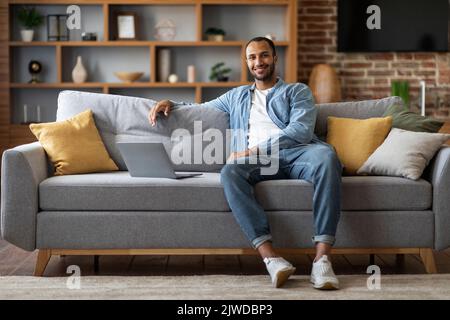 Handsome Black Man Sitting On Couch With Laptop And Smiling At Camera Stock Photo