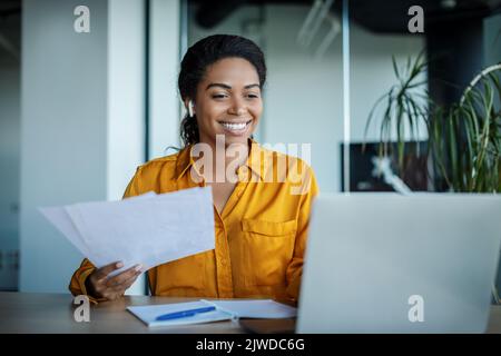 Happy black businesswoman working with papers and having video call on laptop, sitting in office Stock Photo
