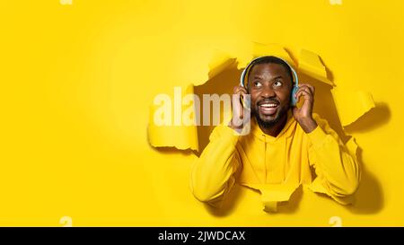 Young happy black man peeking out hole isolated on yellow background and listening to music with headphones Stock Photo